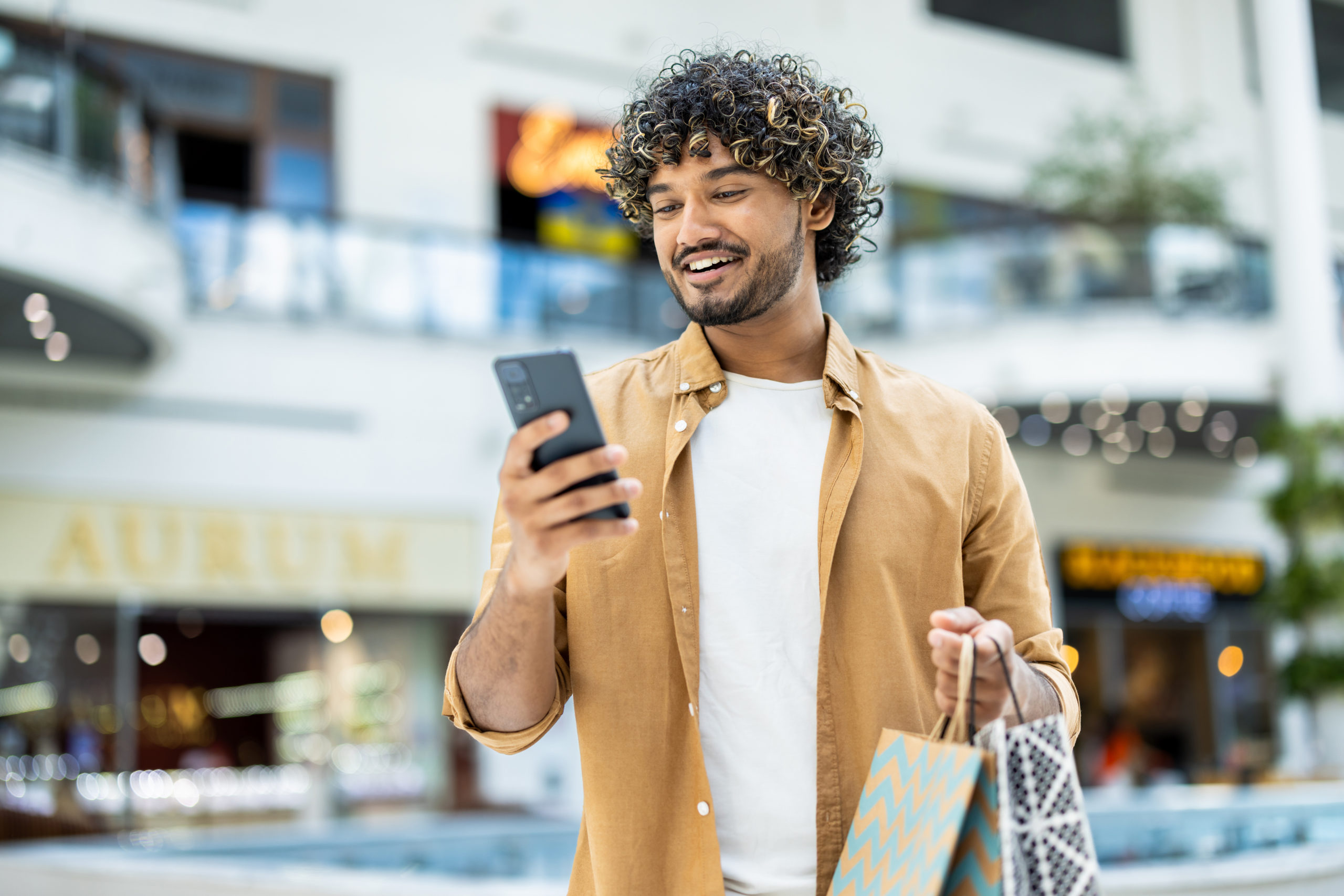 A young man in a mall looks at his phone while carrying shopping bags smiling