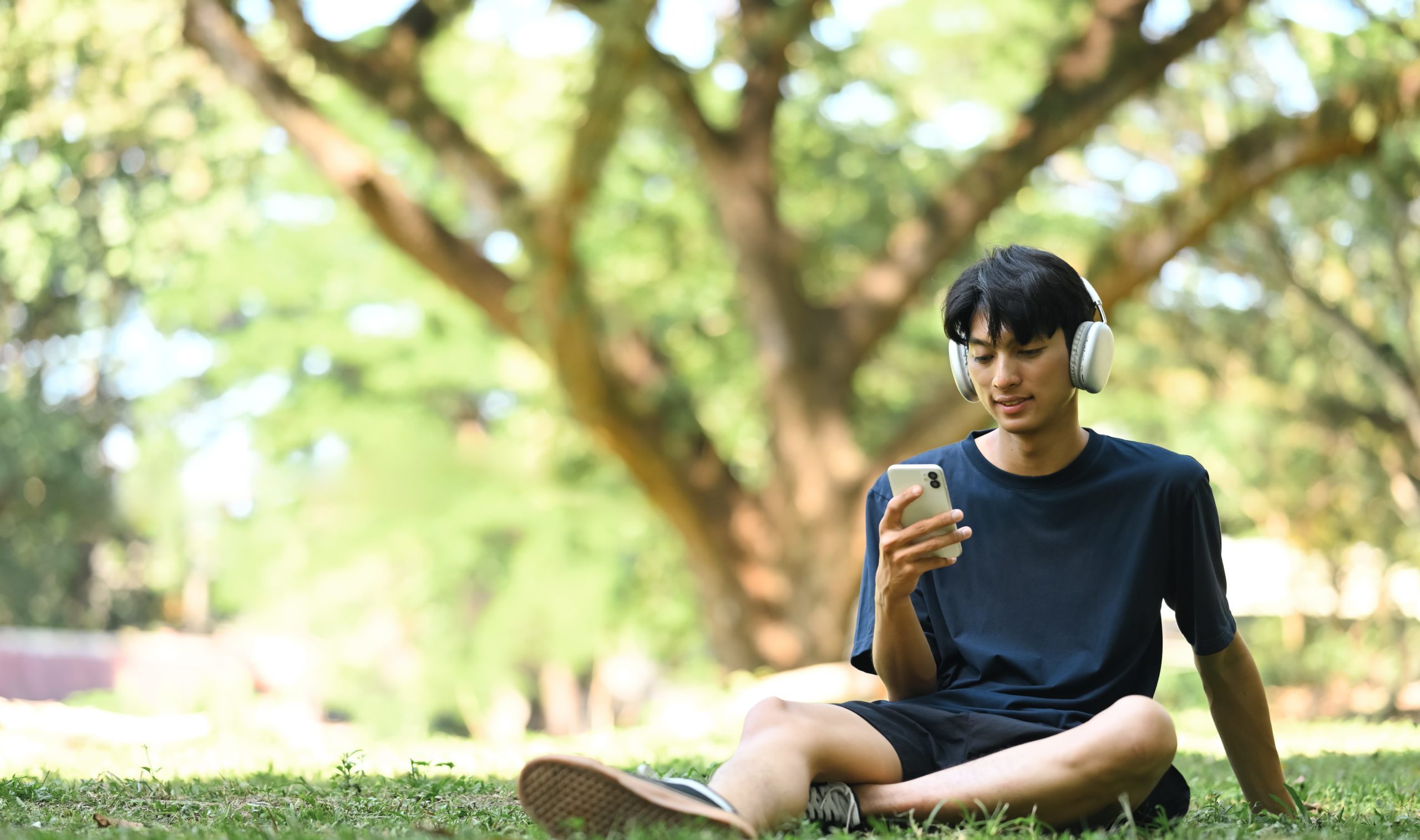 Young man listening to music and using mobile phone on the grass in a sunny park
