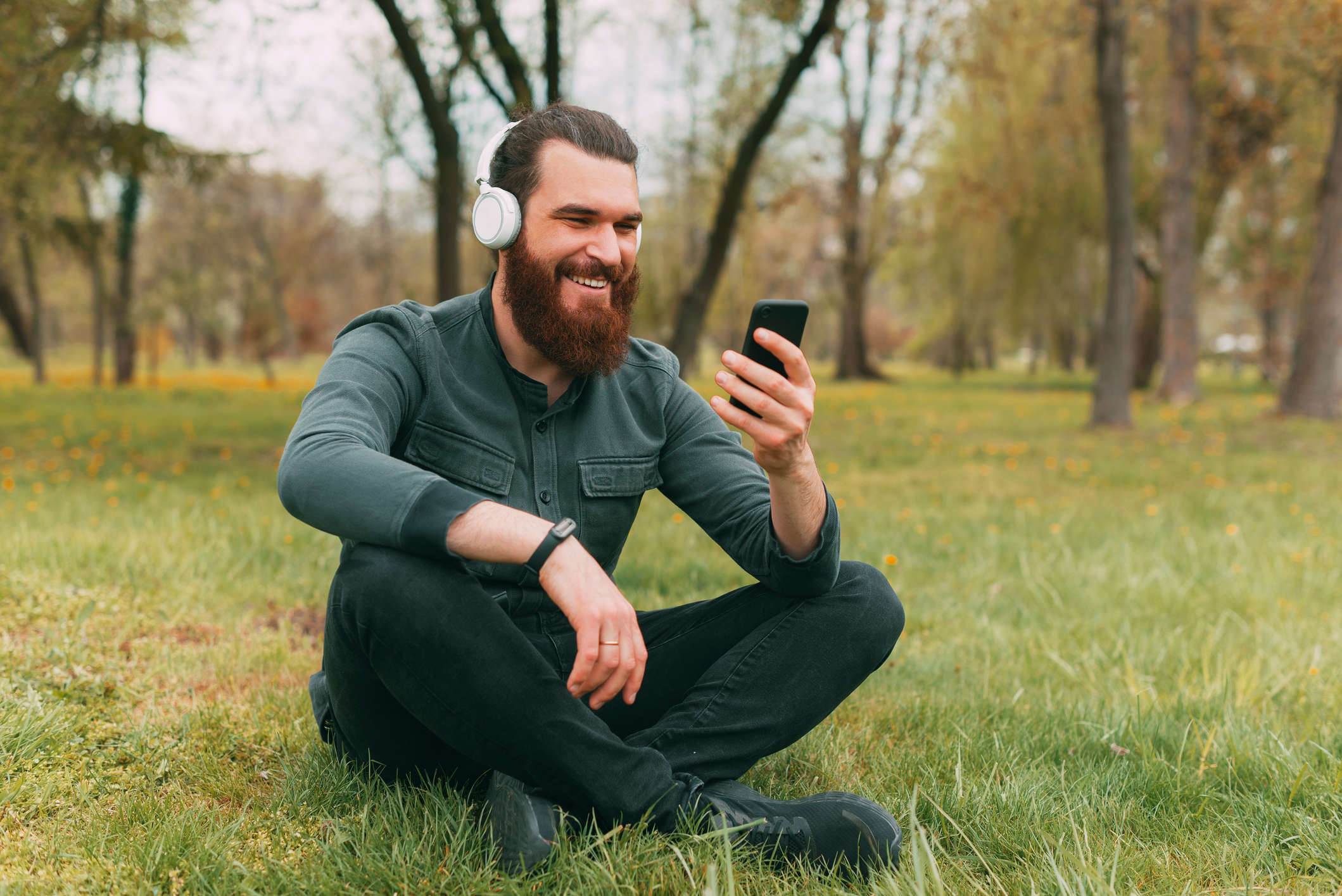 Happy bearded hipster man sitting on grass in park listening music and using smartphone