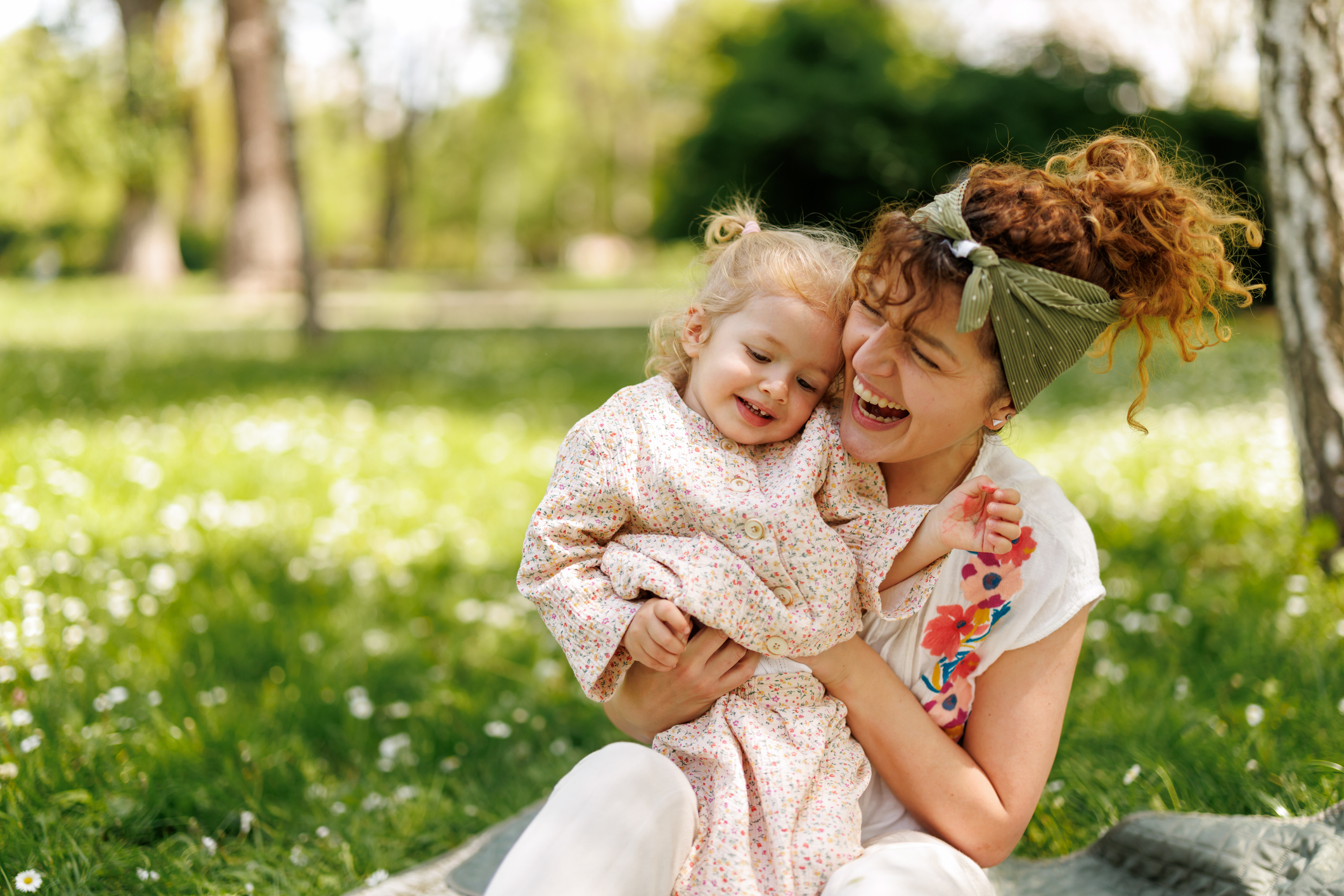 Affectionate mother cuddling and hugging her cute little daughter while sitting on blanket in blooming spring nature