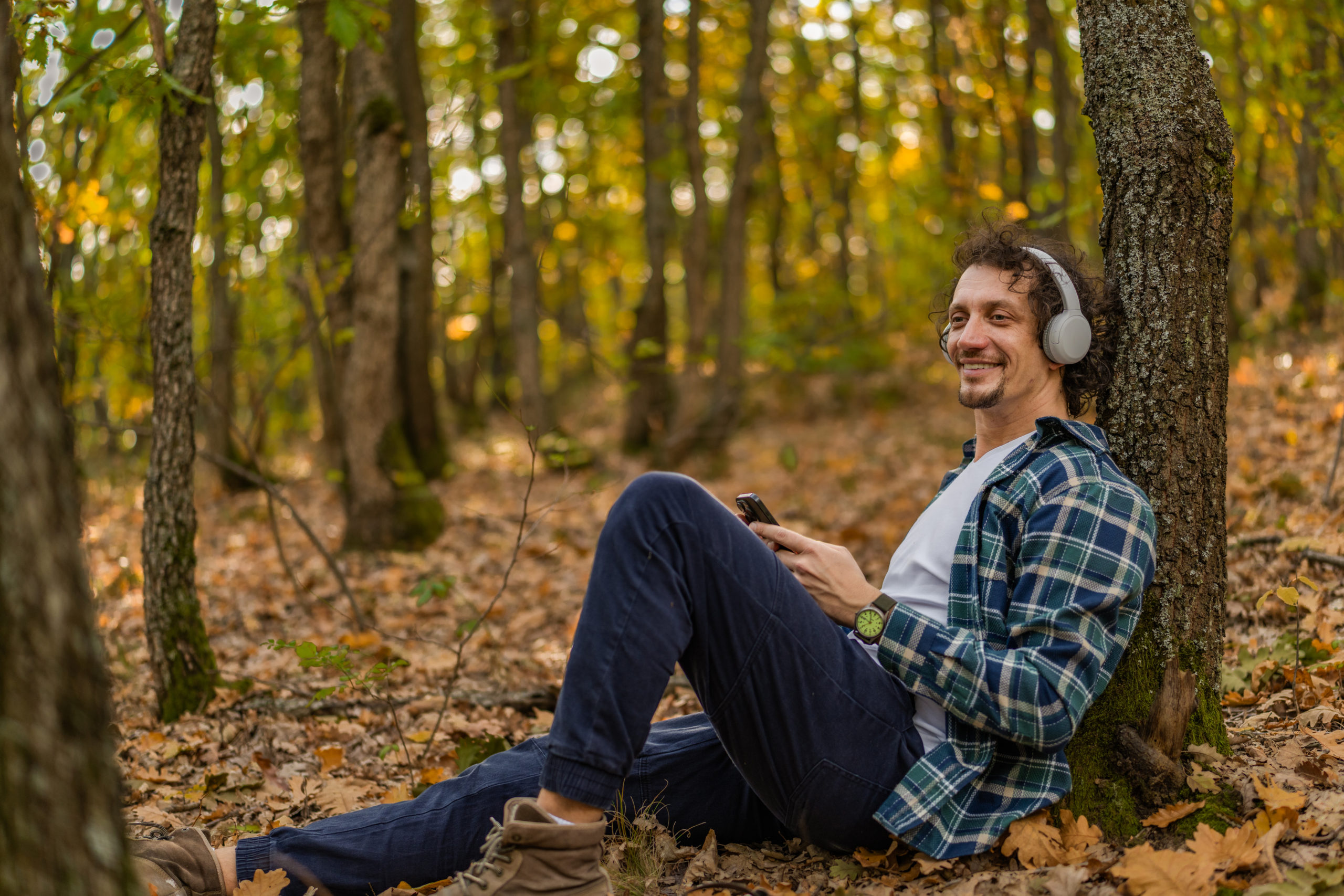 Man with wavy hair resting in nature leaning on a tree trunk listening the music and enjoying in the autumn day